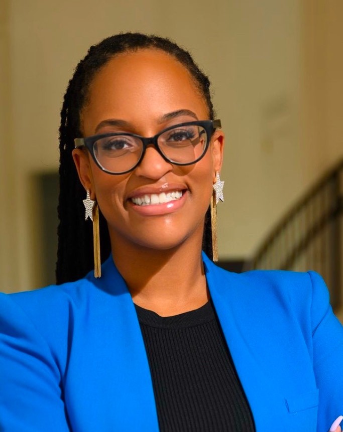 Ashley Sutherland, Co-Founder of She Exhales — a Black woman in a cobalt blue blazer and statement earrings, smiling warmly