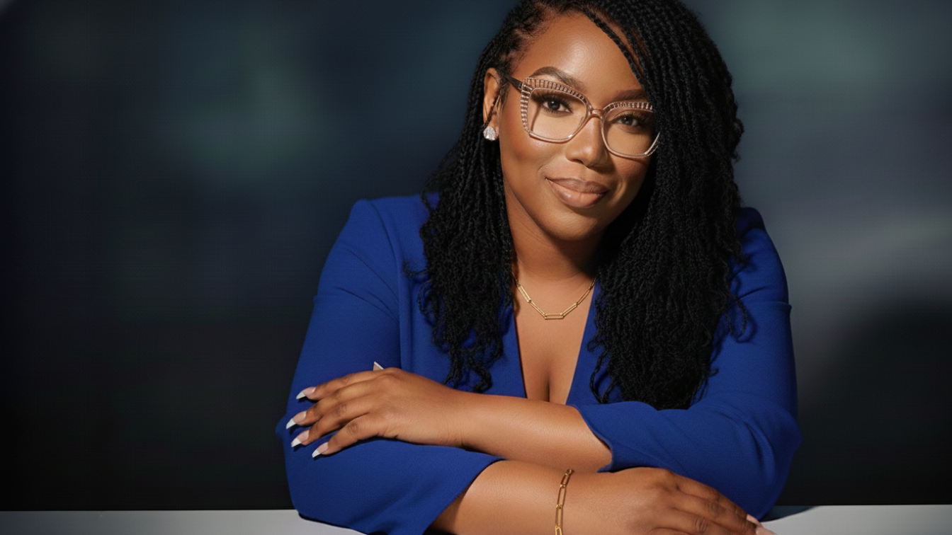 Alisha Phipps, Founder of She Exhales — a Black woman in a royal blue blazer and clear cat-eye glasses, arms crossed, smiling softly at the camera